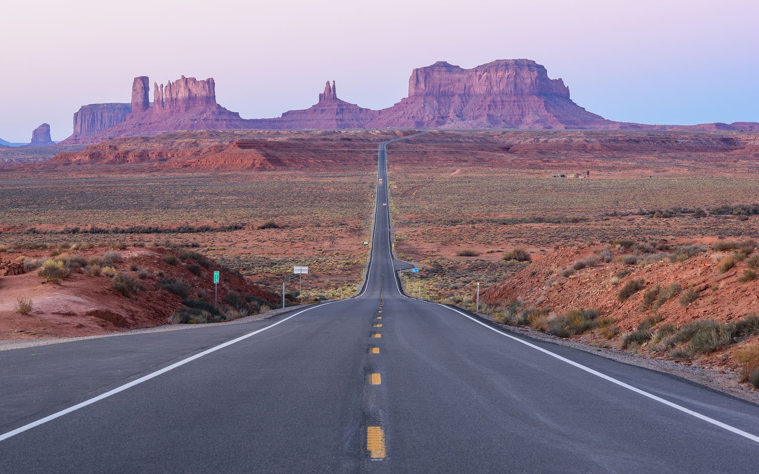 A straight two-lane highway running to the horizon toward the buttes of Monument Valley under a pink and lavender sky